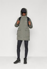 Žalia pūkinė striukė su kapišonu, dviem priekinėmis kišenėmis ir užtrauktuku. Suderinta su juodomis leginsais ir tvirtais juodais žygio batais.