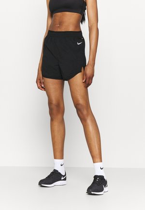 Atletisk person iført sorte Nike-shorts, sort sports-bh, hvide Nike-strømper og sorte Nike løbesko mod en ensfarvet hvid baggrund.