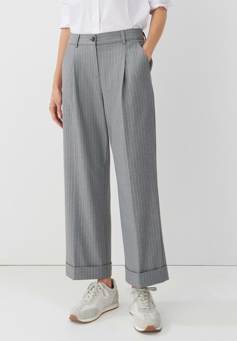 Personne portant un pantalon large à fines rayures gris avec des ourlets retroussés, des baskets blanches et une chemise blanche, debout avec une main dans la poche.