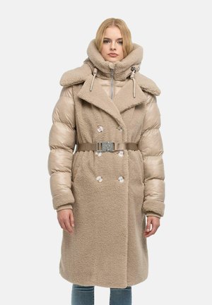 Donna che indossa un cappotto beige con cintura, maniche imbottite e collo alto, in piedi davanti a uno sfondo semplice.