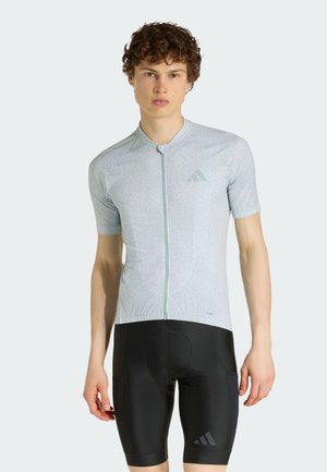 Ung mand iført lysegrå cykeltrøje med korte ærmer og sorte polstrede cykelshorts, stående foran en ensfarvet baggrund.