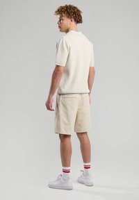 Miestnská svetlobežová polo košeľa s textúrovaným pletením, skombinovaná so svetlo krémovými šortkami. Biele tenisky s červenými pruhovanými ponožkami. Neutrálne pozadie.