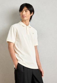 Πόλο μπλουζάκι σε κρέμα βαμβάκι, με κλασικό γιακά, τρεις κουμπάκια, κοντά μανίκια και μια μικρή λογότυπη λεπτομέρεια στο στήθος.