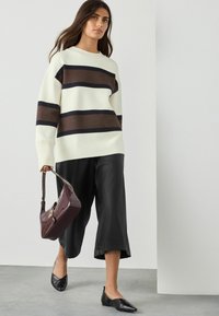 Frau, die drinnen spaziert, trägt einen weißen Pullover mit braunen und schwarzen Streifen, schwarze Culottes, schwarze spitze Ballerinas und hält eine burgunderfarbene Handtasche.