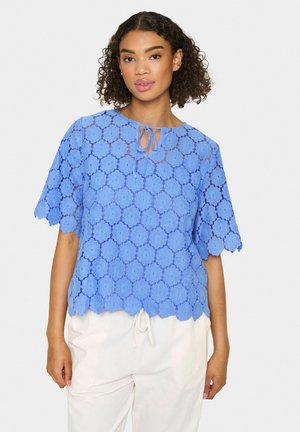 Top in pizzo blu con motivo floreale, maniche corte, bordi ondulati e chiusura a fiocco sul collo. Indossato sopra pantaloni chiari.