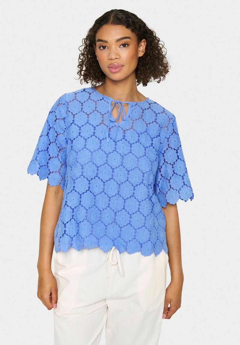 Top in pizzo blu con motivo floreale, maniche corte, bordi ondulati e chiusura a fiocco sul collo. Indossato sopra pantaloni chiari.