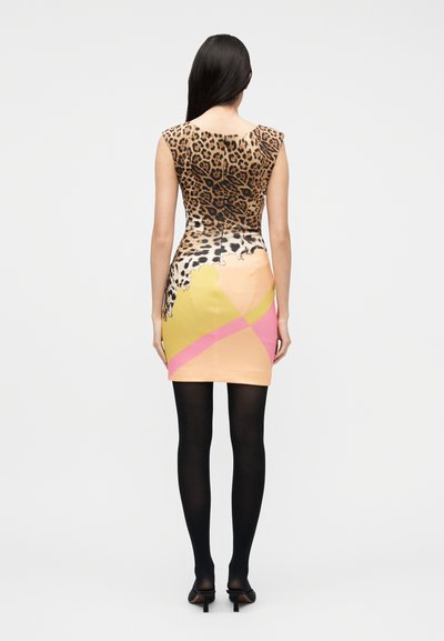Γυναίκα με μακριά σκούρα μαλλιά, που φοράει μίνι φόρεμα χωρίς μανίκια με ζωηρό σχέδιο ζώου και μπλοκ χρωμάτων, μαύρες καλσόν και μαύρα τακούνια, στραμμένη με την πλάτη.