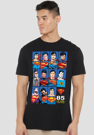Junger Mann trägt ein schwarzes T-Shirt mit einem bunten Raster aus zwölf Superman-Comic-Porträts und dem Text "85 Years" unten.