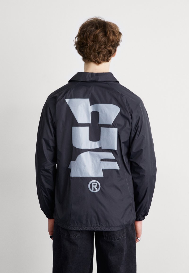 Mustat nailonitakki, jossa on harmaa graafinen logo selässä, kauluksella ja elastisilla hihansuilla. Sileä pinta, rento muotoilu.