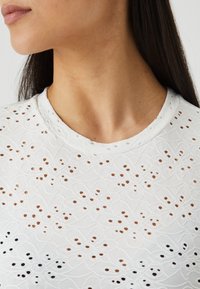Bílý top s detailním výšivkovým designem s květinovým vzorem, kulatým výstřihem a texturovaným povrchem pro prodyšné nošení.