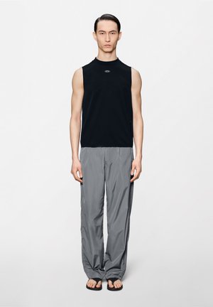 Λεπτός άνδρας μοντέλο στέκεται όρθιος φορώντας μαύρο αμάνικο τοπ, χαλαρά γκρίζα παντελόνια και μαύρα σαγιονάρες, μπροστά σε απλό λευκό φόντο.