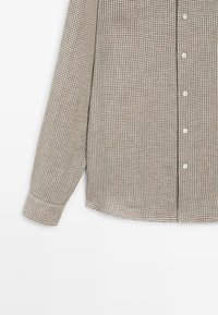 Brązowo-beżowa koszula w kratę z długim rękawem i białymi guzikami, prezentowana na jednolitym białym tle, ukazująca prawą połowę tułowia i rękawa.