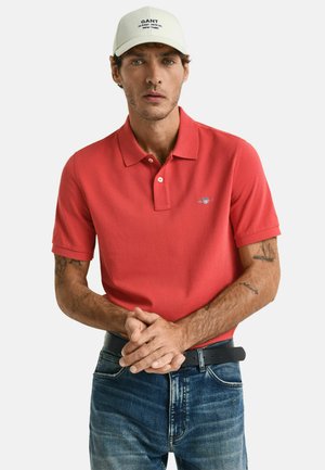 Camisa polo roja de manga corta con cuello. Presenta un logo en el pecho. Combinada con vaqueros de mezclilla azules y un cinturón negro.