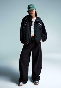 Zwarte nylon jas met witte accenten, witte crop top, loose zwarte broek en groene pet met "geen drama." Zilveren en bruine schoenen.