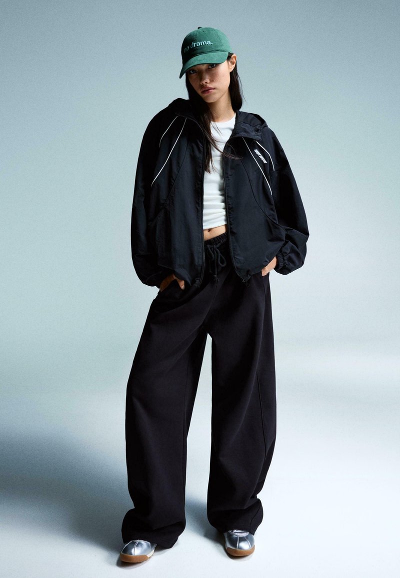 Zwarte nylon jas met witte accenten, witte crop top, loose zwarte broek en groene pet met "geen drama." Zilveren en bruine schoenen.