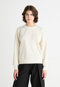 Kremfarget sweatshirt med rund hals, med ribbestrikkede mansjetter og hem, med teksturdetaljer rundt halslinjen. Båret med mørke bukser.