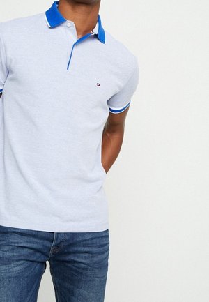 Helles graues Polohemd mit einem blauen Kragen und Ärmelausschnitten. Verfügt über ein kleines Logo auf der linken Brust. Getragen mit blauen Jeans.