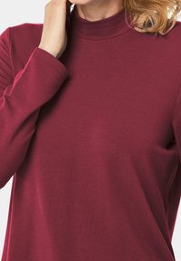 Burgunderfarbener, mock-turtleneck Shirt aus weichem, dehnbarem Stoff. Mit langen Ärmeln und einem lockeren Schnitt, versehen mit dezenten Stichdetails.
