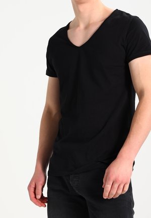 Młoda osoba ubrana w dopasowany czarny T-shirt z dekoltem w serek i czarne spodnie, stojąca z ramionami swobodnie opuszczonymi wzdłuż boków.