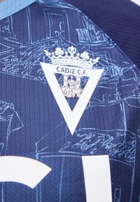 Μπλε αθλητική φανέλα με άσπλο λογότυπο Cadiz C.F., που δείχνει ένα στεφανωμένο τρίγωνο και λεπτομερή γραμμική τέχνη κτηρίων στο φόντο.