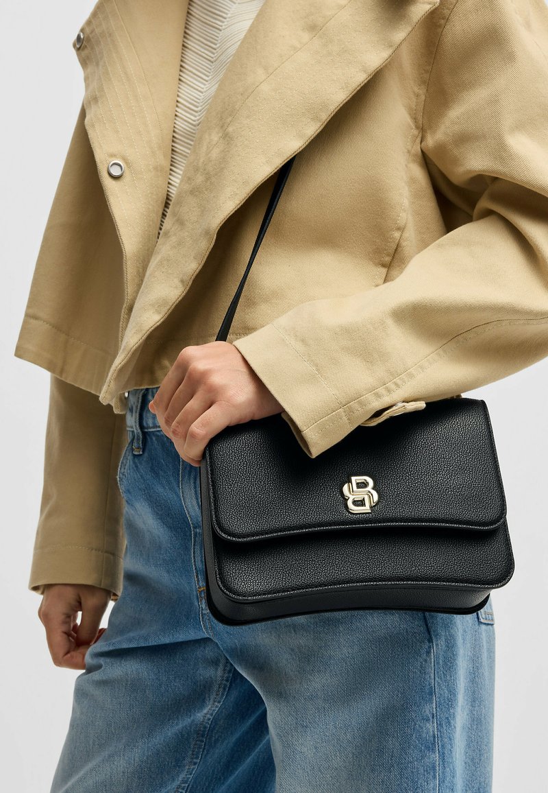 Borsa a tracolla in pelle nera dal design strutturato, con due scomparti e un accento in oro del logo, portata con una tracolla nera su una giacca beige.