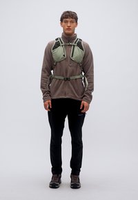 Ελαφρύ πράσινο γιλέκο ενυδάτωσης με ρυθμιζόμενους ιμάντες, φορεμένο πάνω από γκρι φλις μπουφάν, σε συνδυασμό με μαύρο παντελόνι και καφέ παπούτσια πεζοπορίας.