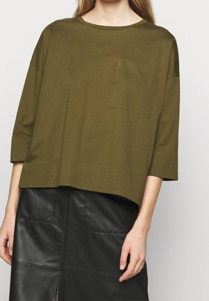 Frau trägt ein olivgrünes, übergroßes Oberteil mit Brusttasche und Dreiviertelärmeln, kombiniert mit einem schwarzen Lederminirock mit Frontschlitz.