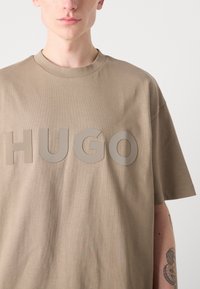 Bēša kokvilnas T-krekls ar brīvu piegriezumu, ar lielu, reljefīgu "HUGO" logo gaišākā tonī un īsām piedurknēm.
