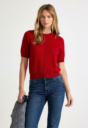 Kvinde med langt blond hår iført en rød kortærmet knappet sweater og blå jeans, der holder en sort-hvid ternet jakke.