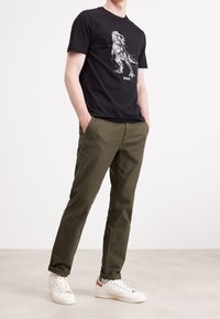 Czarny T-shirt z białym wzorem, zestawiony z oliwkowozielonymi spodniami i białymi trampkami. Model stoi z rękami w kieszeniach.