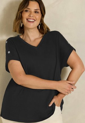 Smilende kvinde med kort hår bærer en sort V-hals skjorte med knapdetaljer på ærmerne og poserer med den ene arm bøjet.
