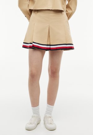 Persona que lleva puesta una minifalda plisada beige con el dobladillo a rayas en azul marino, blanco y rojo, combinada con calcetines blancos y zapatillas blancas, de pie frente a un fondo blanco.