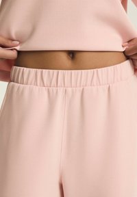 Osoba podnosząca jasnoróżową bluzkę, odsłaniając środek ciała i elastyczny pas dopasowanych jasnoróżowych spodni.