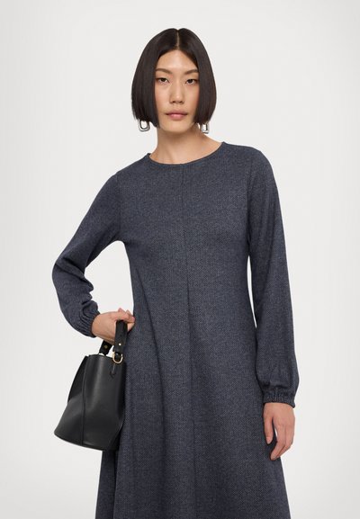 Abito a zigzag blu navy con maniche lunghe a sbuffo e scollatura rotonda, abbinato a una piccola borsa a secchiello in pelle nera con dettagli dorati.