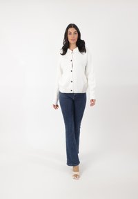 Cárdigan blanco de punto con botones de madera, tela texturada y puños acanalados, combinado con vaqueros oscuros de corte acampanado y sandalias claras.