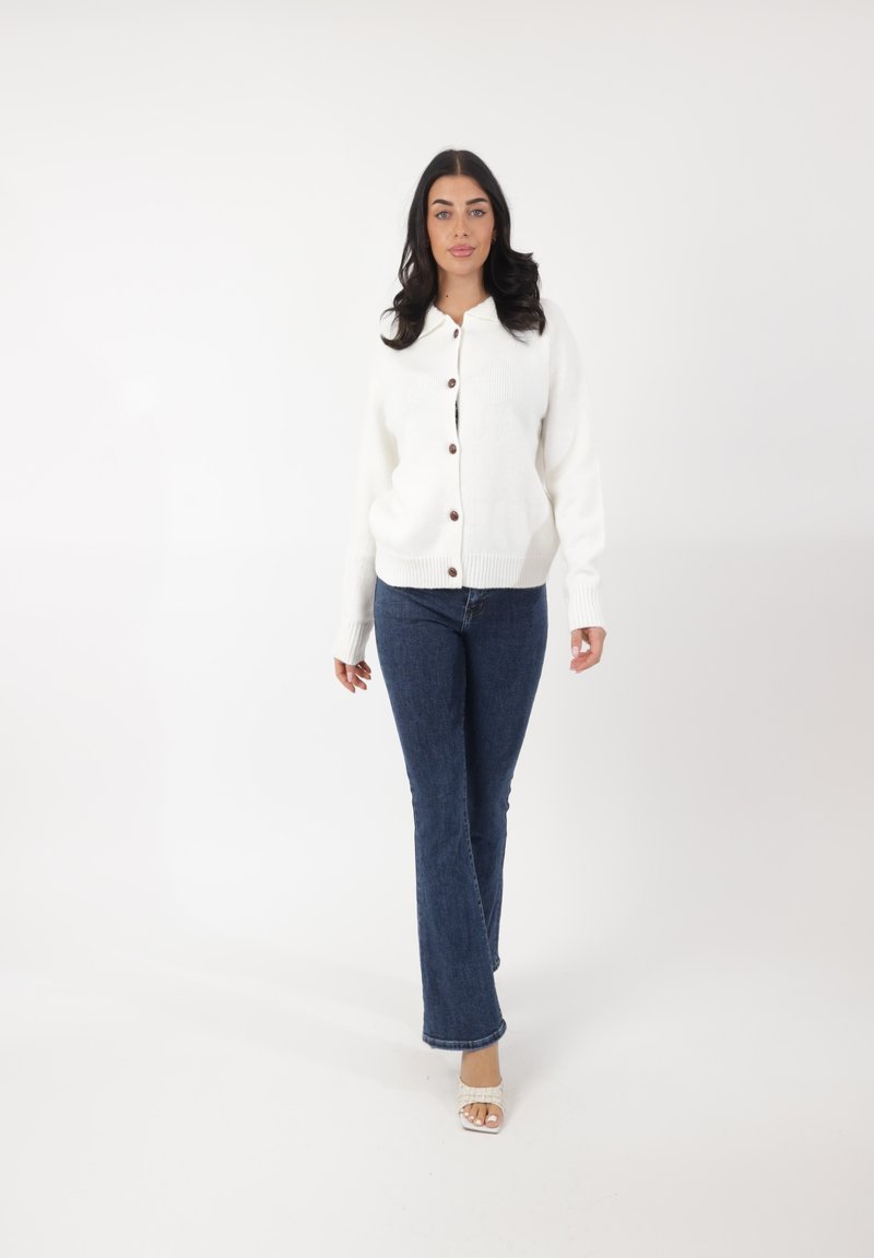 Cárdigan blanco de punto con botones de madera, tela texturada y puños acanalados, combinado con vaqueros oscuros de corte acampanado y sandalias claras.