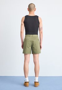 Μαύρο ριγέ αμάνικο μπλουζάκι και ελαιόχρωμες βερμούδες με δύο πίσω τσέπες. Το άτομο έχει κοντά μαλλιά και φοράει λευκές κάλτσες και καφέ παπούτσια.