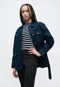 Veste bleu marine avec des boutons dorés, plusieurs poches avant, associée à un chemisier rayé noir et blanc et un pantalon noir ajusté.