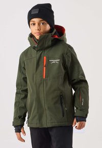 Grön utejacka med huva, med orange detaljer, dragkedjeförsedda fickor och en logotyp, bärs med en svart mössa. Slät, slitstark tyg.