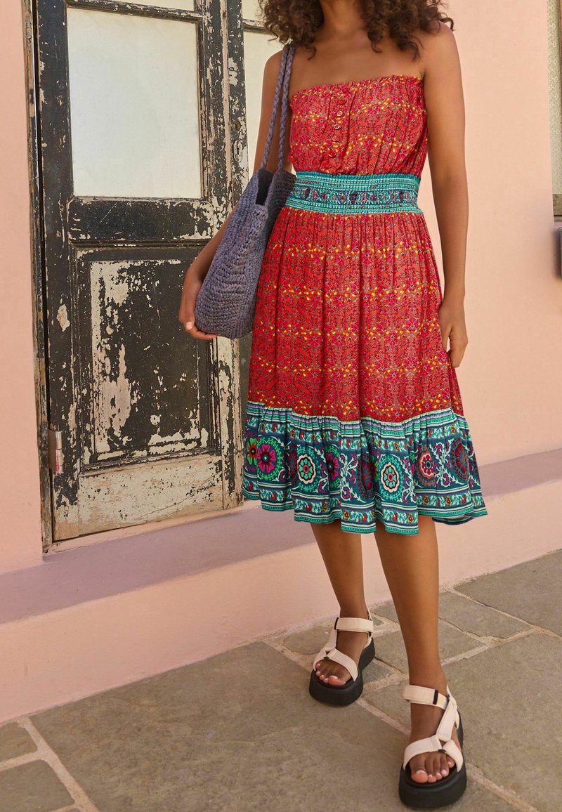 Femme en robe bustier rouge à motifs avec bordure florale bleu sarcelle, debout tenant un sac cabas gris tissé près d'une porte en bois abîmée.