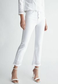 Jeans acampanados de denim blanco con recortes de ojales en el dobladillo. Presenta una cintura ajustada con cierre de botón y un acabado texturizado y suave.