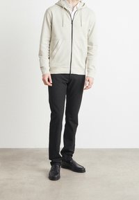 Felpa grigia chiaro con zip, cappuccio e tasche frontali, abbinata a pantaloni neri slim-fit e scarpe di pelle nere. Tessuto strutturato visibile.