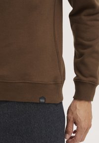 Brun sweatshirt med ribbet mansjetter og hem, med et lite logo-merke. Stoffet ser mykt og strukturert ut, kombinert med mørke rutete bukser.