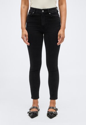 Vedere frontală a unei persoane care poartă blugi negri skinny cu talie înaltă și pantofi negri cu vârf ascuțit și două curele cu catarame.