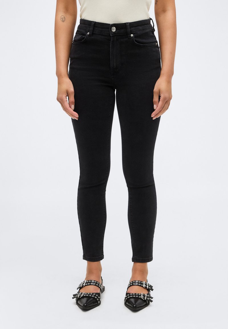 Εμπρόσθια όψη ενός ατόμου που φοράει μαύρο στενό τζιν ψηλής μέσης και μαύρα μυτερά παπούτσια με διπλές λουρίδες και αγκράφες.