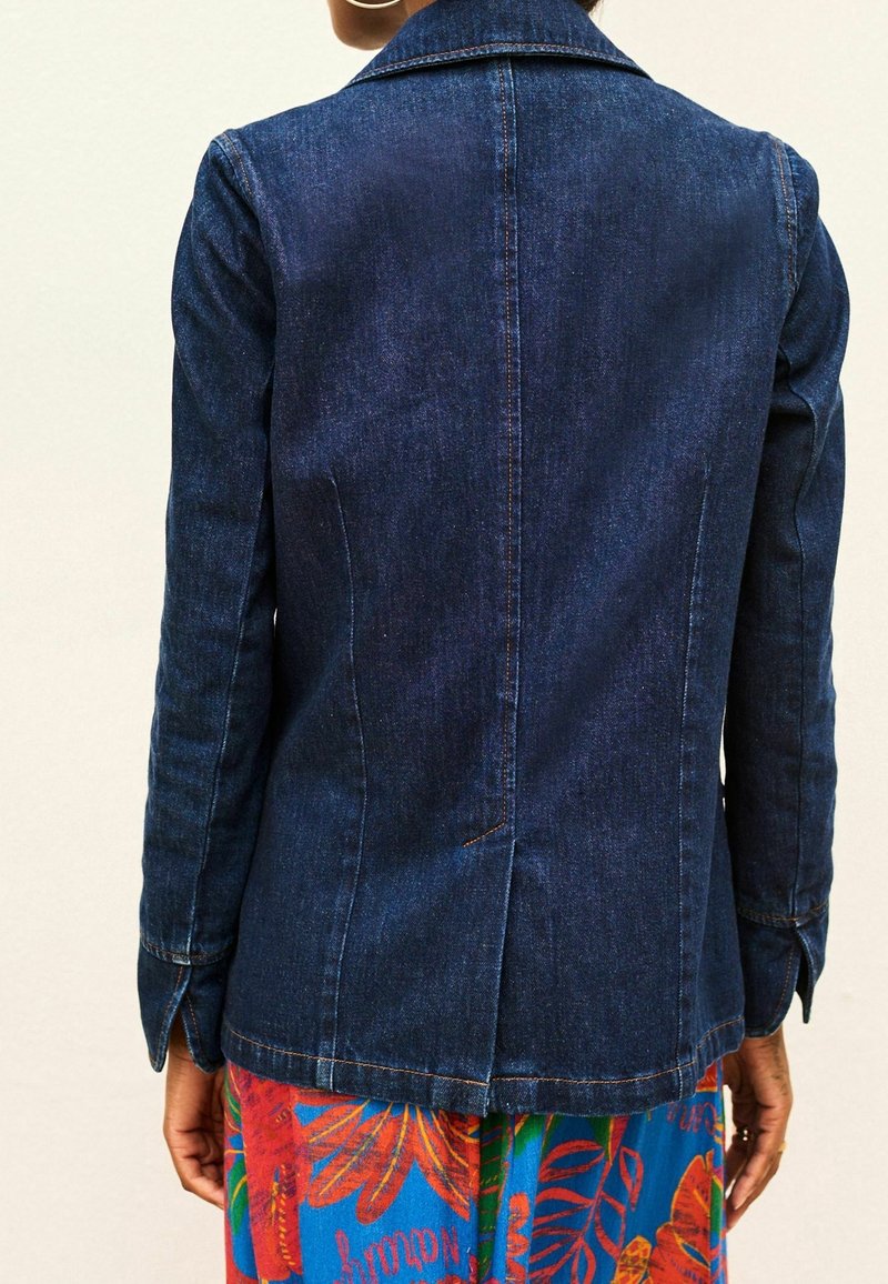 Personne portant une veste en jean bleu foncé avec des surpiqûres oranges, debout de dos, vêtue d'une tenue colorée avec des motifs rouges et bleus.