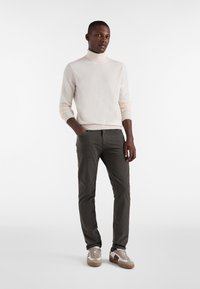 Moški stoji z eno roko v žepu, oblečen v kremno polo majico, temne oprijete hlače in bež superge pred enobarvno belo podlago.