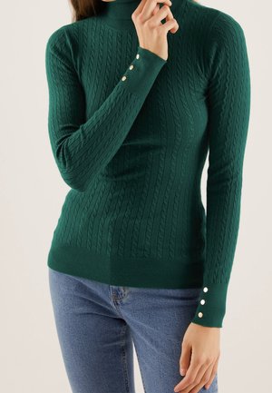 Femme portant un pull ajusté en maille torsadée vert foncé avec des boutons dorés sur les poignets et un jean bleu, posant avec une main près du visage.