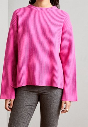 Vrouw draagt een felroze geribbelde trui met wijde mouwen en grijze broek met visgraatpatroon, staand tegen een effen achtergrond.