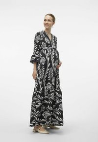 Robe maxi noire avec des motifs floraux et de feuilles blancs, décolleté en V, manches cloche 3/4 et jupe avec volants. Associée à des chaussures beiges à enfiler.